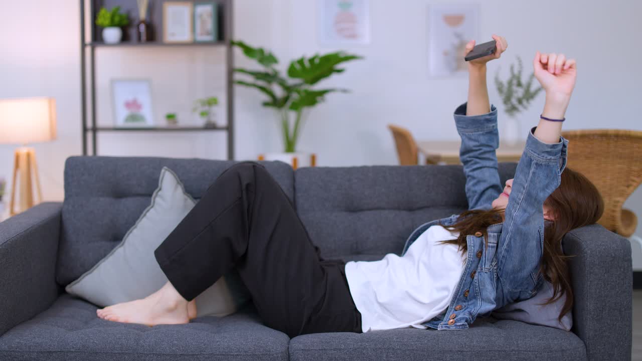 Una giovane donna con una giacca di jeans si rilassa su un divano mentre usa il suo smartphone