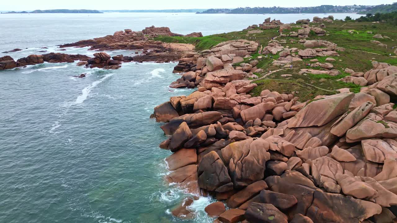 Aerial drone shot flying over unique rock formations of Pink Granite Coast, rugged coastline, hiking trails with tourists, Brittany, France