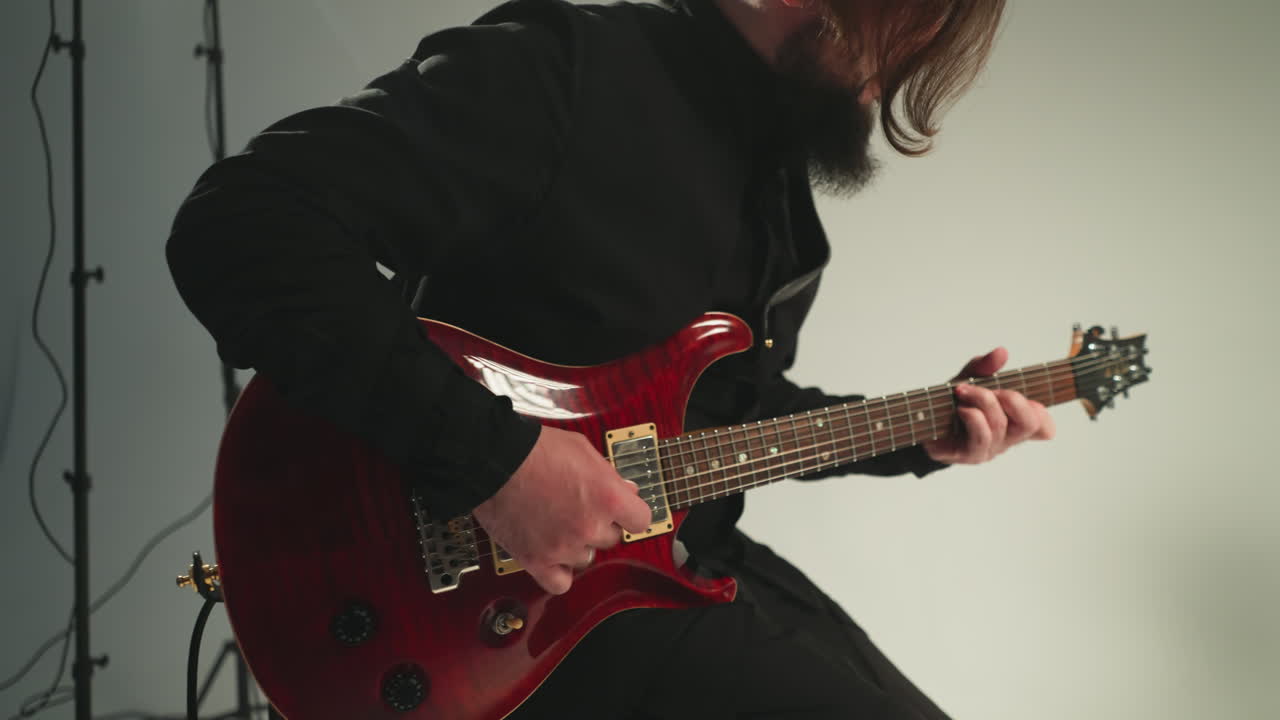un guitarrista con un traje negro se sienta y toca una guitarra eléctrica roja contra un fondo gris, con otra guitarra visible en el fondo. la cámara hace zoom