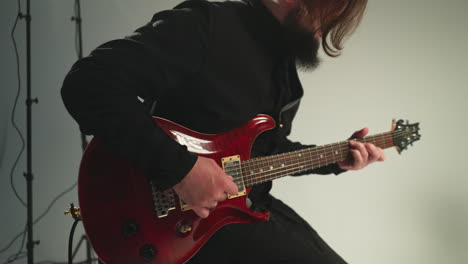 a guitarist in a black outfit sits and plays a red electric guitar against a grey background, with another guitar visible in the background.the camera zooms out