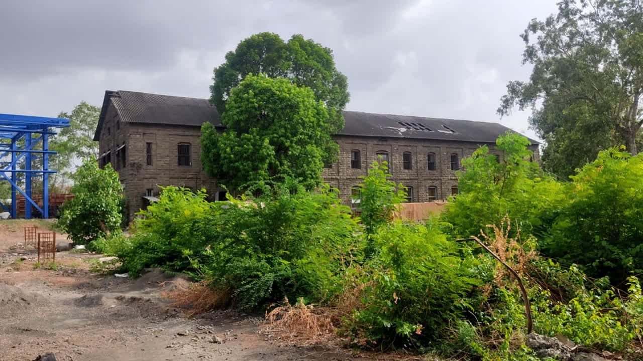 Antiguo edificio de piedra de dos plantas con un tejado dañado y varias ventanas, parcialmente oculto por densos árboles y arbustos verdes.