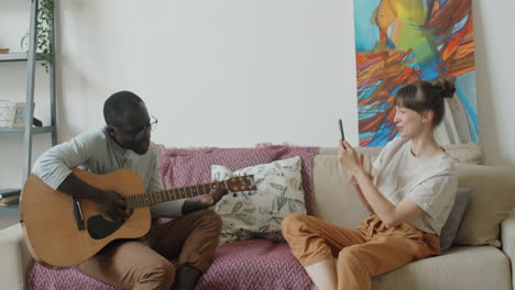 african american man playing guitar and singing as wife filming video with phone