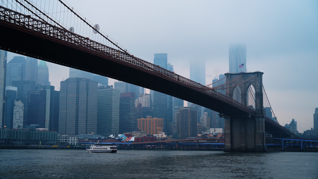 Brooklyn Bridges steintårn og nedre Manhattan skyline i tåke. Utsikt fra vannet som viser Brooklyn Bridges steinpylon, en forbipasserende ferge og tåkebelagte skyskrapere
