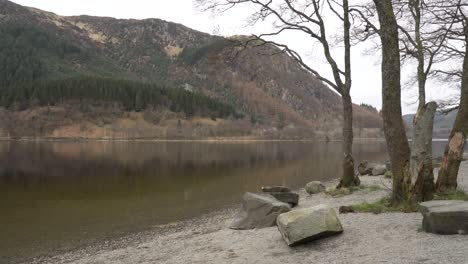 Toma-De-Seguimiento-Y-Panorámica-Desde-La-Costa-De-Un-Lago-Con-Rocas-En-Primer-Plano-Y-Montañas-En-El-Fondo-En-Un-Día-Nublado-En-Escocia