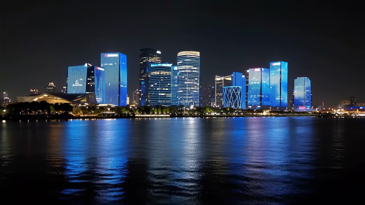 Modern towers remain steady along the distant waterfront while gentle ripples shift blue and white reflections across foreground water, framed in a wide nighttime city view.