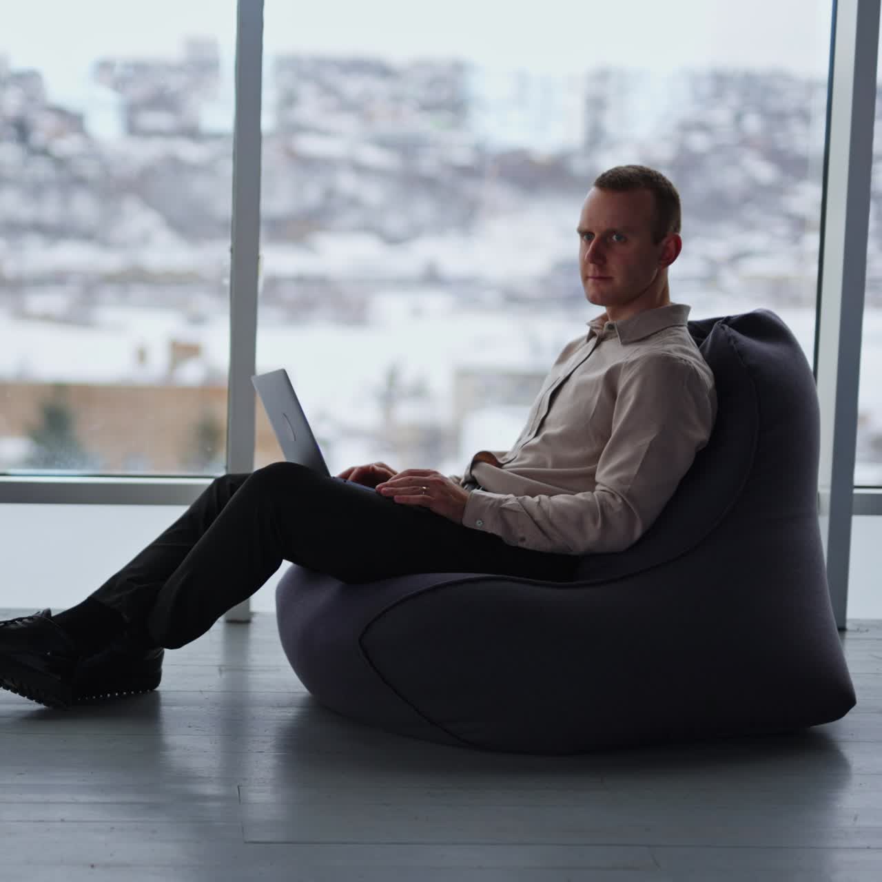 Middle-aged male works on laptop sitting in bean bag chair. Man distracts and looks aside, putting hand to his forehead. Cityscape in blur at backdrop