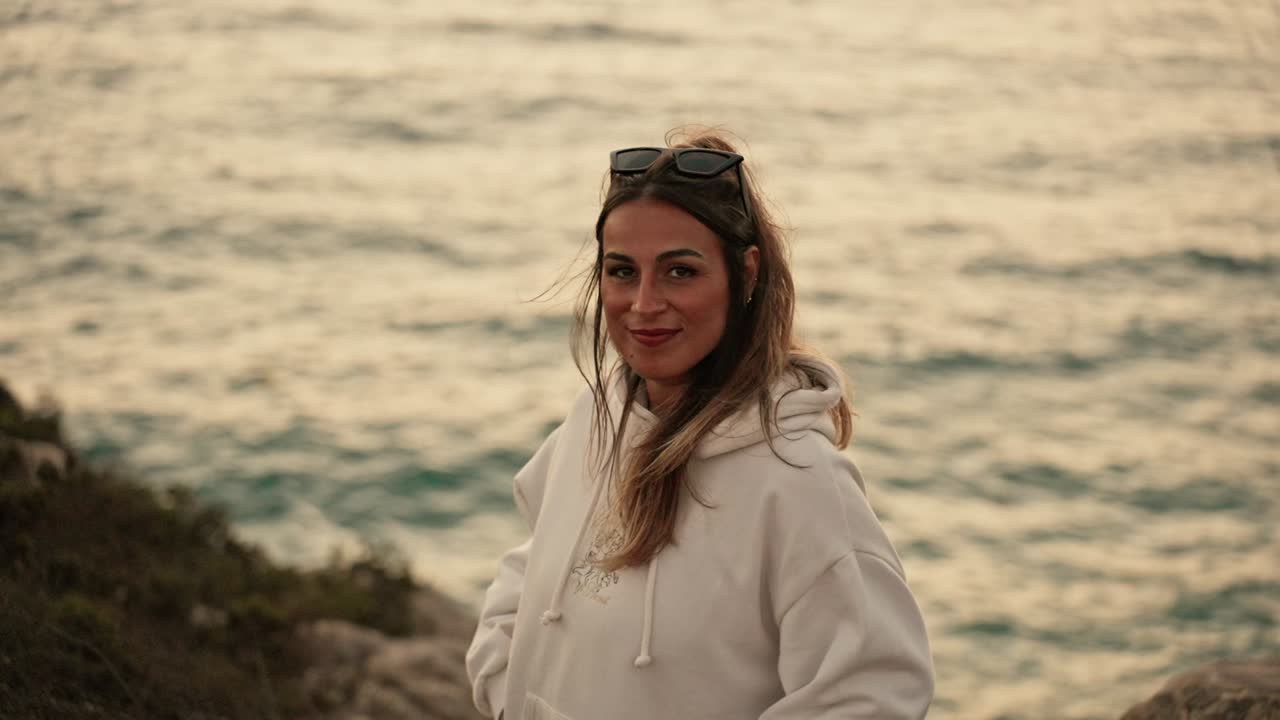 Attraktiv spansk kvinde i boheme-stil smiler genert ved havet under solnedgang