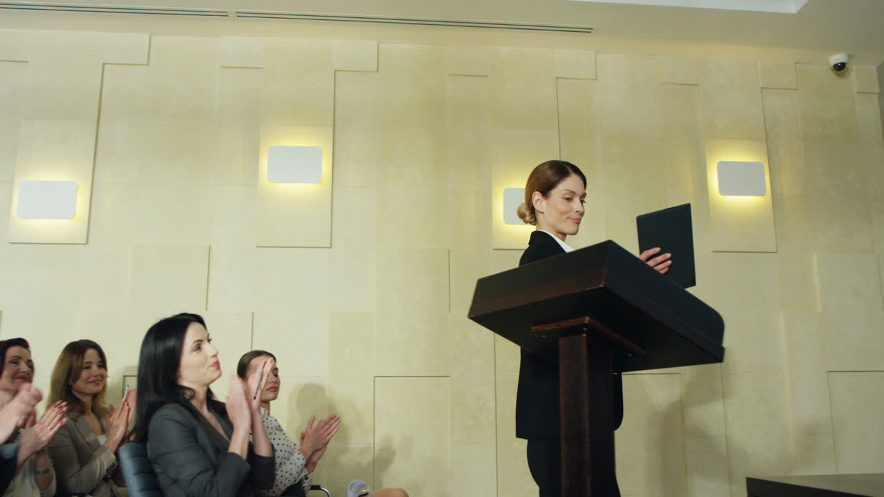 사람들이 박수를 치는 동안 백인 젊은 여성 사업가가 회의실에 들어서고 연단에 올라옵니다.