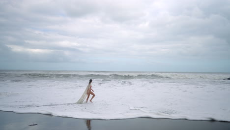 woman in bikini and long white cover walks slowly along a moody, cloudy beach