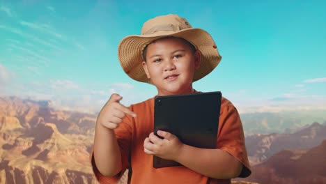 asian boy with a hat and binoculars smiling and pointing to a tablet while traveling at the top of mountain. boy researcher examines something, travel tourism adventure concept, close up