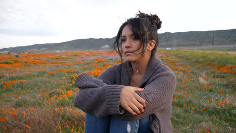 a cute and attractive girl looking sad and serious sitting alone in a rural field of wild flowers with wind blowing her hair at dusk slow motion