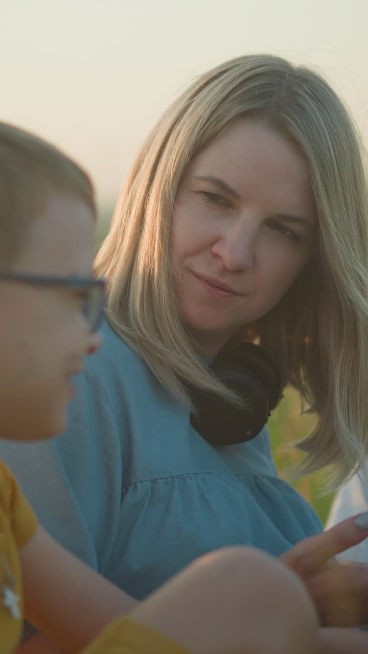 una madre alegre en un vestido azul multitarea en una computadora portátil y teléfono en un campo cubierto de hierba, involucrándose con su hijo de amarillo, que ríe mientras su otro hijo de blanco se sienta en silencio a su lado