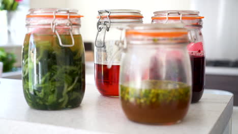 handheld shot of fermented wild garlic, fermented red berries, pickled wild garlic flowers and raspberry vinegar in jars in a kitchen with the chef working in the kitchen in the blurred background