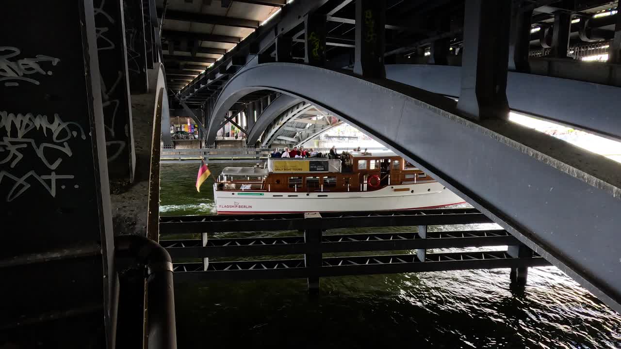 Tour boat passes under steel urban bridge with graffiti, cityscape, and natural daylight riverside view
