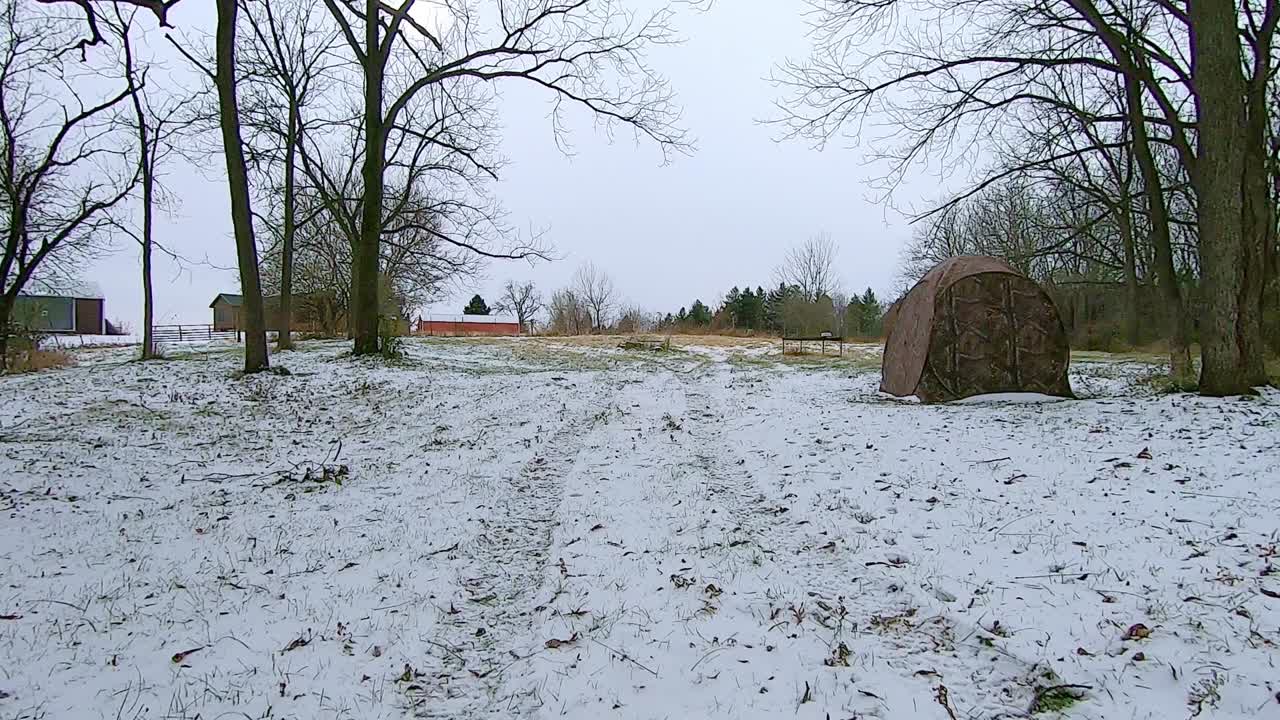 All terrain vehicle ATV while driving on a snow covered trail through a grassy field near timber on an early winter afternoon; a barn, farm house and deer stand are in the distance; point of view