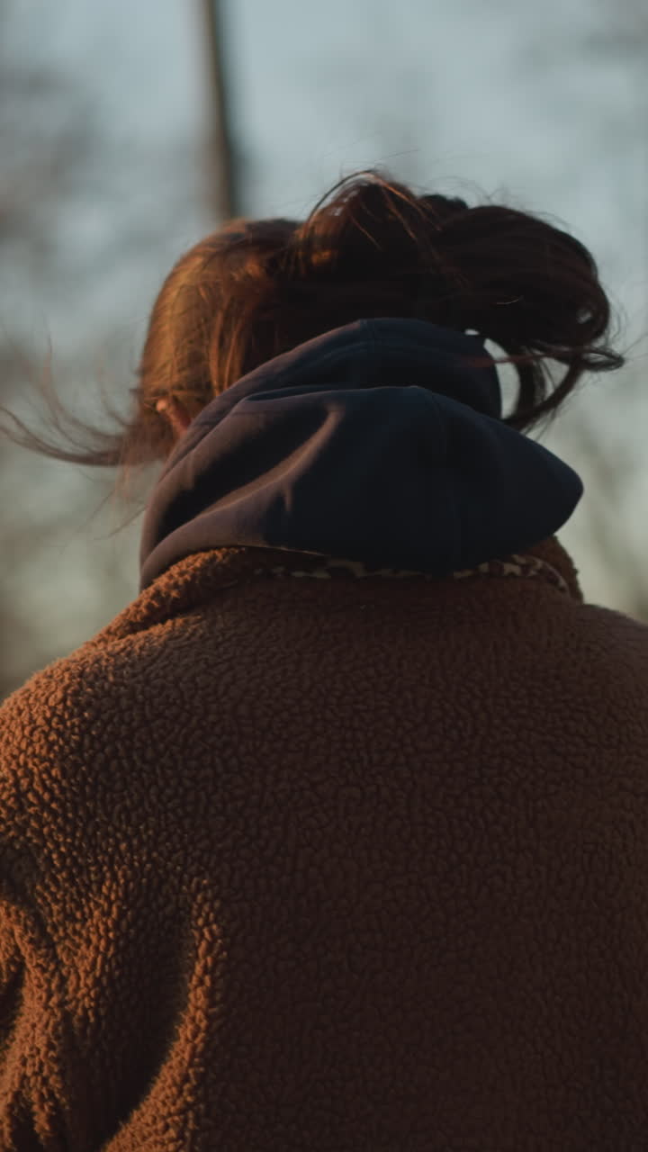 A somber girl walking through a park, with the wind gently blowing her hair. She is dressed in a brown coat and a hoodie, her expression reflecting deep thought