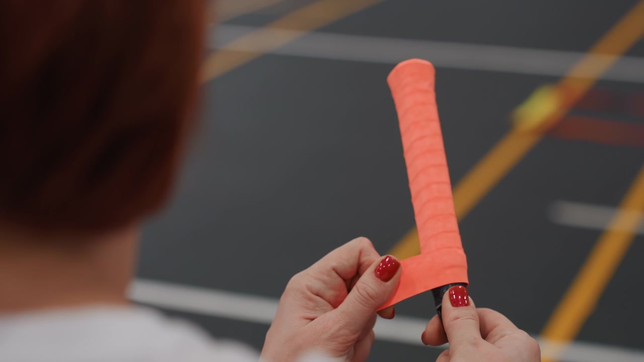 Primo piano di una donna che fissa un nastro adesivo colorato sull'impugnatura di una racchetta da badminton su un campo al coperto, giocatrice amatoriale che regola la tensione e l'allineamento, atmosfera di riscaldamento veloce prima della partita con linee del campo ben visibili.