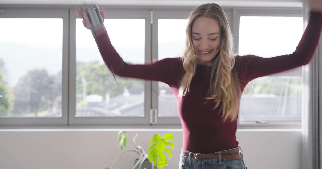 Mujer bailando música del teléfono con auriculares con cable, girando en un dormitorio luminoso y vistiendo una blusa granate.