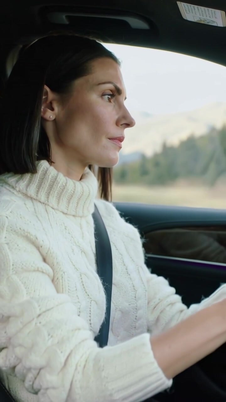Video verticale: Donna alla guida con un maglione color crema su una strada rurale, che regola il volante, con la cintura di sicurezza allacciata.