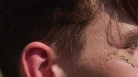 close ear chin view of white man facing riverbank, mud camouflage under eye, hand adjusting headband, sunlit outdoor setting with grass and water blur in background, expression of focused relief
