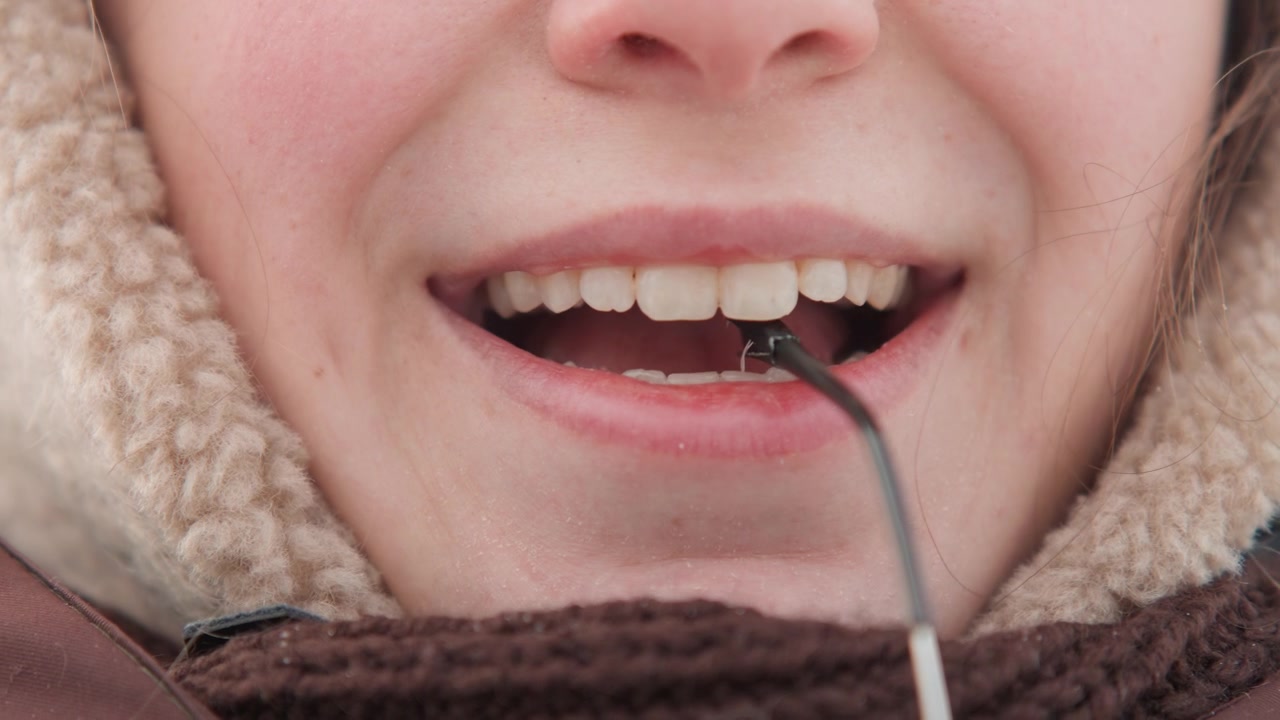 Center-framed close-up shows lower face of young adult outdoors in winter gently bringing thin metal probe toward slightly open mouth, touching lower lip and visible teeth with neutral focused