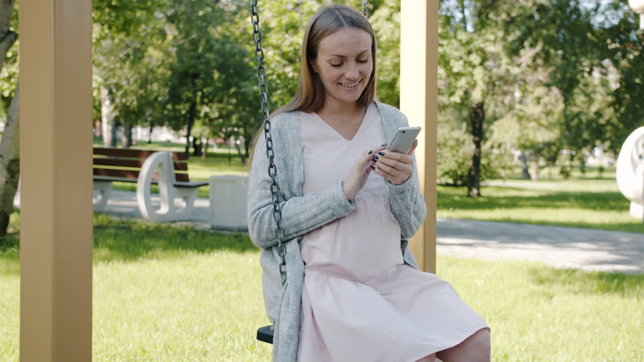 Pregnant woman using phone on a swing in a park