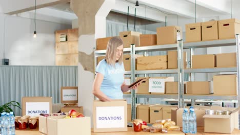 caucasian woman volunteer packing donation boxes with food and water while typing on the tablet in charity warehouse