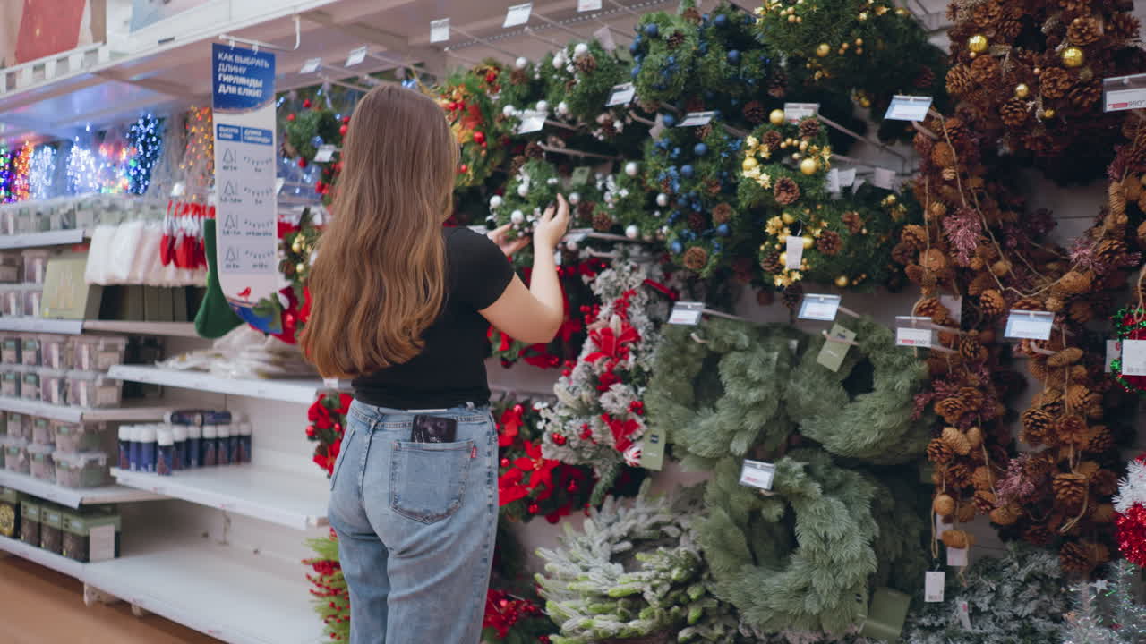 vista trasera de una mujer revisando las decoraciones festivas en una tienda minorista, examinando coronas, guirnaldas y adornos navideños, estantes llenos de luces brillantes y coloridas decoraciones estacionales