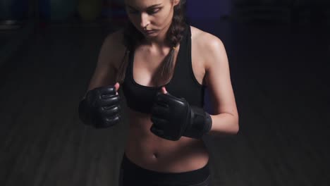 Woman-taking-on-boxing-gloves-on-hands-with-black-boxing-wraps-in-dark-room.-Slow-Motion-shot