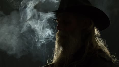 closeup dark portrait: backlit cowboy with long beard smokes cigarette