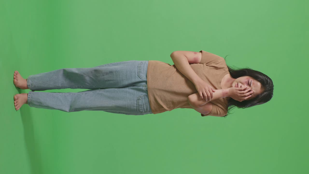 폭력의 희생자인 젊은 아시아 여성의 몸이 얼굴과 팔에 상처를 입은 이 가득 차 있으며, 그녀의 상처받은 눈은 녹색 스크린 배경 스튜디오에서 울고 있습니다.