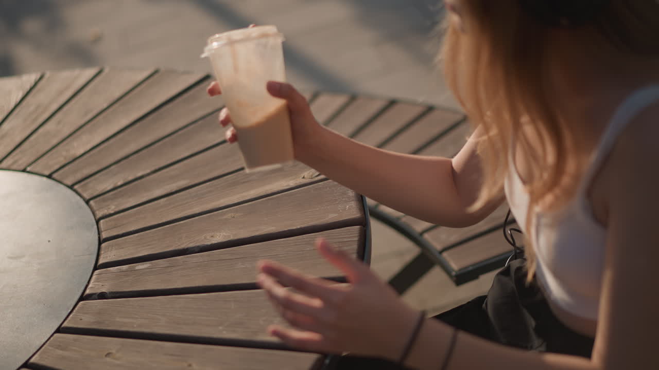 Donna bionda con capelli bianchi seduta all'aperto al tavolino di un bar in legno, con cuffie e top senza maniche, ascolta la musica mentre gesticola e rovescia scherzosamente un latte macchiato freddo, estate soleggiata