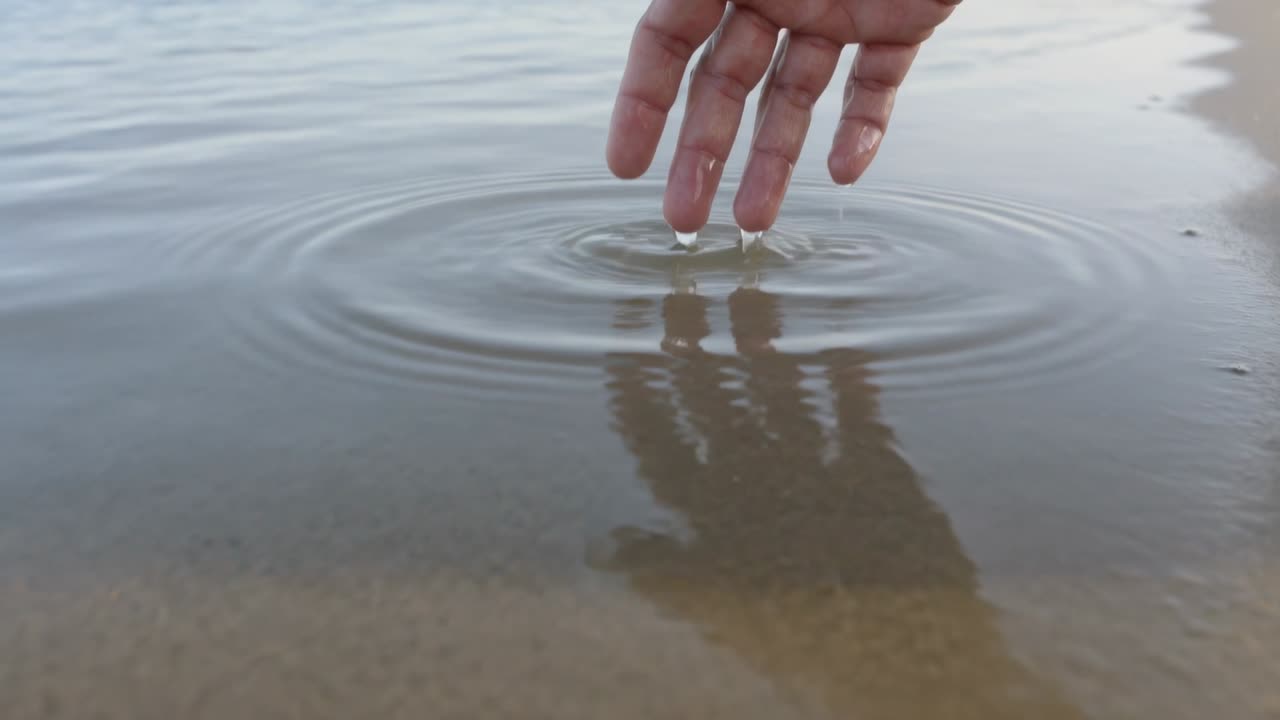 Abbassando la mano destra con un anello di metallo, le punte delle dita sfiorano l'acqua della riva per creare increspature.