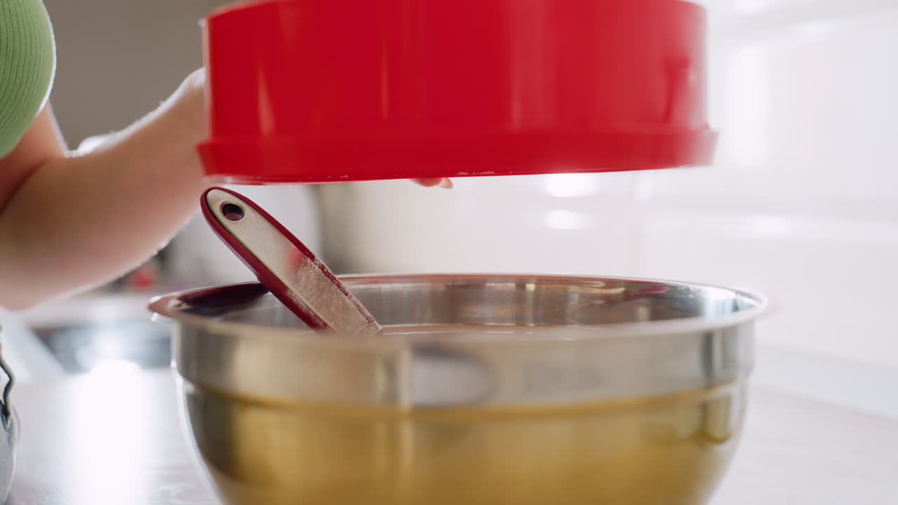 Close up of chef in green top using red sifter to filter flour into stainless bowl during baking preparation, flour dust falling smoothly while mixing utensil rests inside bowl on kitchen counter