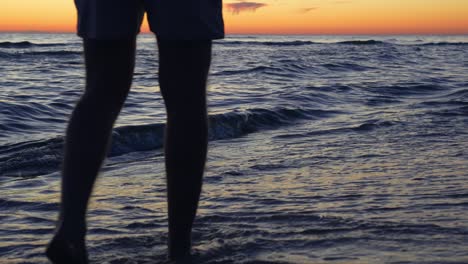 Vista-Trasera-De-Las-Piernas-De-Un-Joven-Caminando-Por-La-Orilla-Del-Mar-Al-Atardecer,-Cámara-Lenta