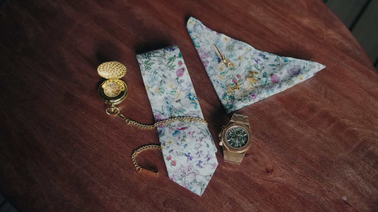 Orologio da tasca d'oro, gemelli, orologio da polso e pochette floreali posati su legno.