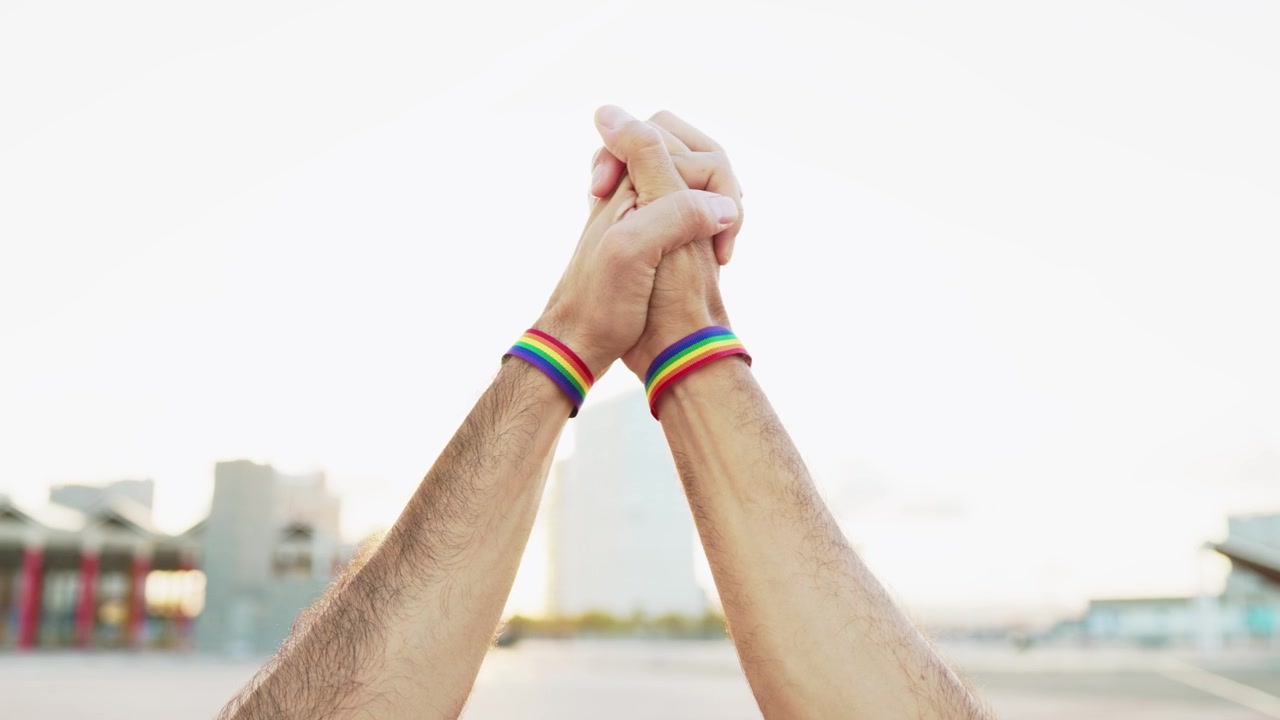 Primo piano di mani con braccialetti Pride arcobaleno che si tengono.