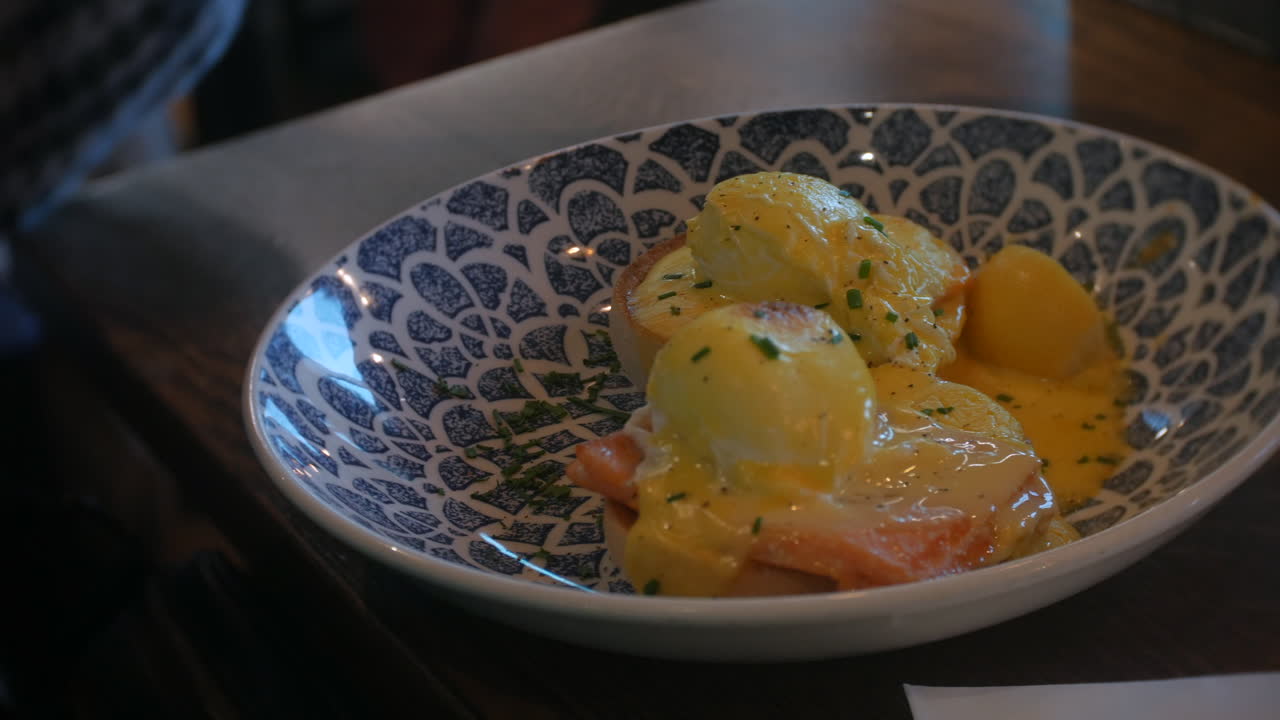 En vakkert anrettet Eggs Benedict med hollandaisesaus og gressløk på et utsmykket blått og hvitt fat, perfekt for eksklusive restaurantmenyer eller luksuriøs frokostmerkevarebygging