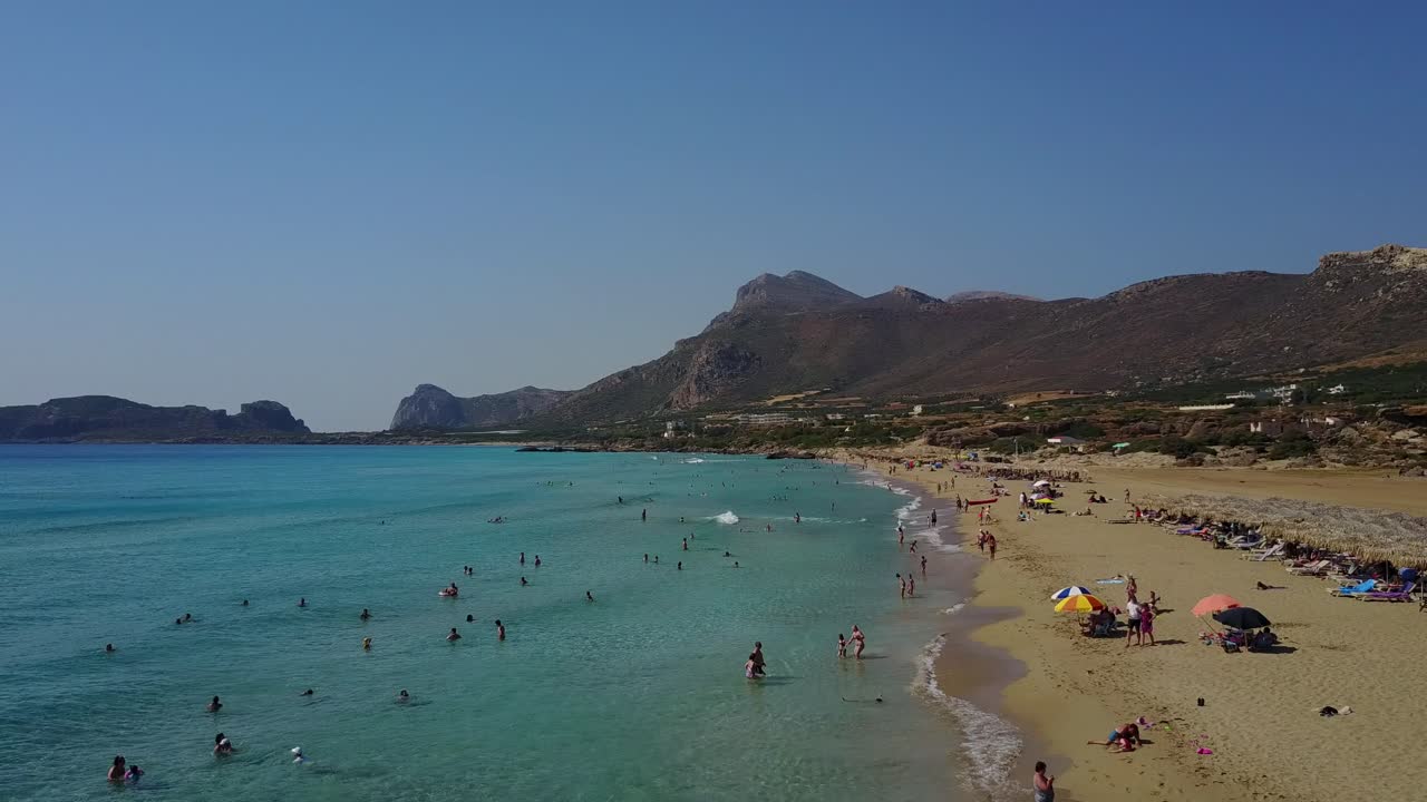 Aerial view of white sand Falassama beach with clear water, tourists, and mountain ranges in Crete, Greece