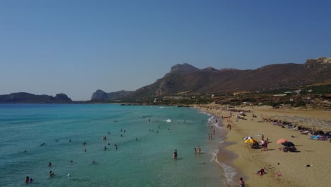 Aerial-view-of-white-sand-Falassama-beach-with-clear-water,-tourists,-and-mountain-ranges-in-Crete,-Greece