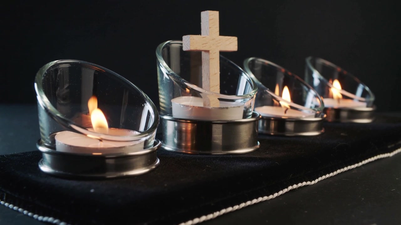 La cámara se mueve, enfocando una cruz de madera sobre 4 portavelas de té de cristal en una almohadilla de terciopelo negro.