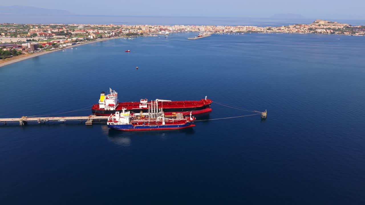 Drone orbiting two oil tankers docked at offshore platform near Sicilian refinery with bay and blue sea visible