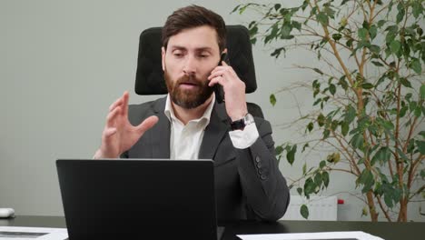 concentrated stylish businessman working on laptop computer in office interior. male worker answering on phone call. man talking on smartphone at remote workplace. business man calling phone indoor.