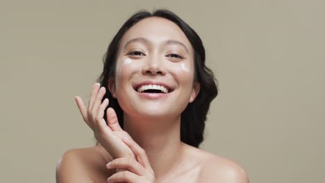 어두운 머리카락을 가진 행복한 아시아 여성이 베이지색 배경에 얼굴에 크림을 바르며, 느린 움직임
