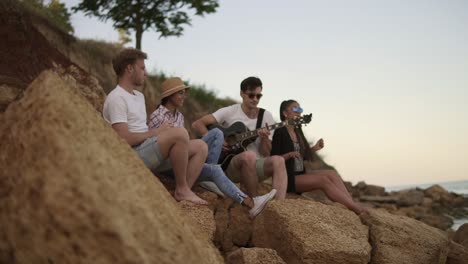 Group-of-young-cheerful-people-sitting-on-the-rocks-by-the-seashore-and-playing-guitar,-singing-songs-and-dancing.-Slow-Motion