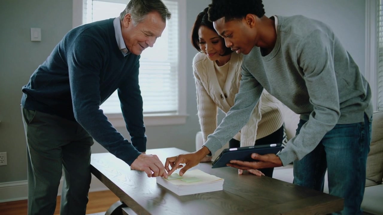 Placing sticky note, three colleagues leaning over dining table inspecting papers consulting tablet