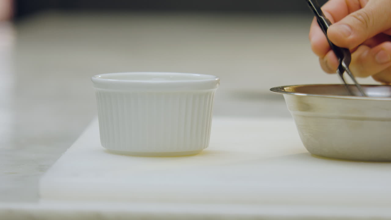 Close-up of a chef's hand using chef's tongs to place apple slices in a white ceramic mold, next to a metal container on a white chopping board