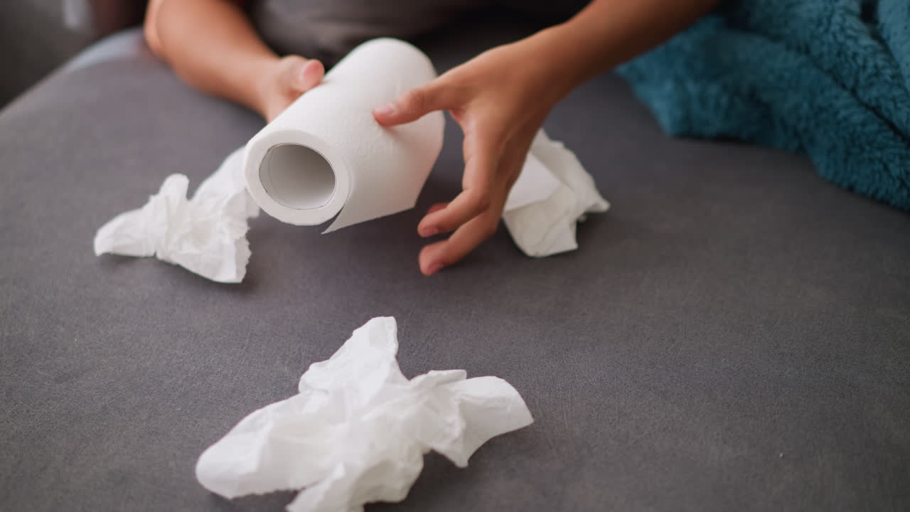 Close-up of hand reaching for tissue roll as used tissues lie on couch, sick person recovering at home, using tissues, with clutter around, and comfort of being at home