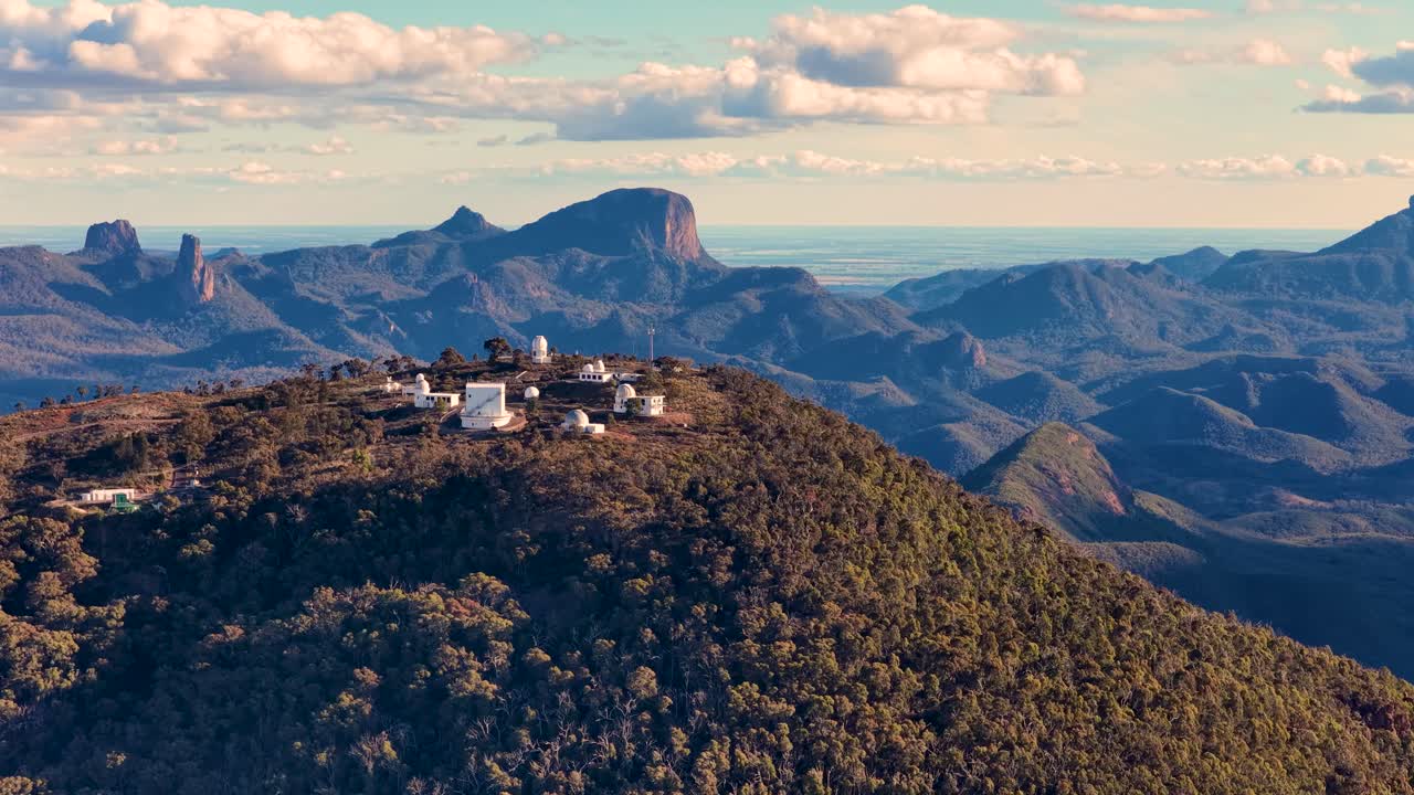 Aerial footage circles a mountaintop observatory complex in Warrumbungle National Park, Australia, with dramatic sunset lighting and expansive mountain views
