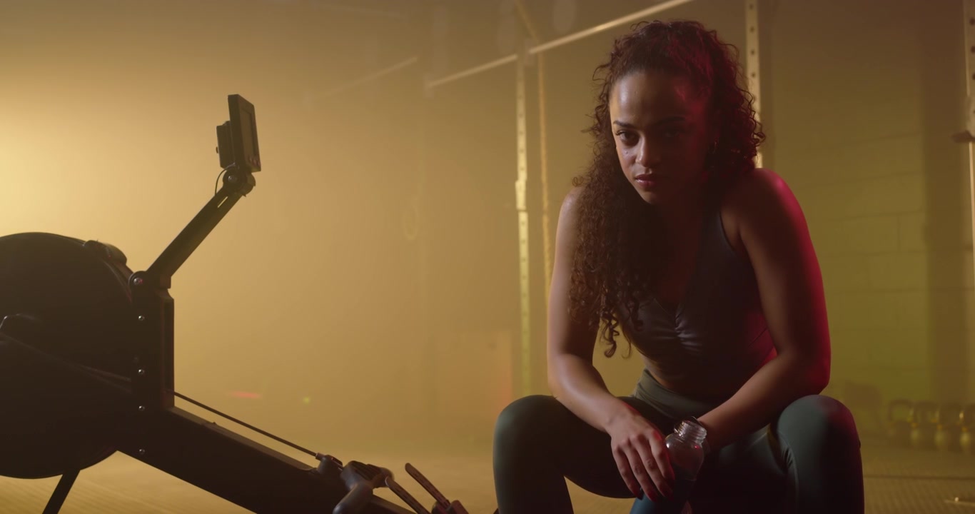 Woman in sportswear sitting on rower after interval, drinking bottle and checking watch and phone